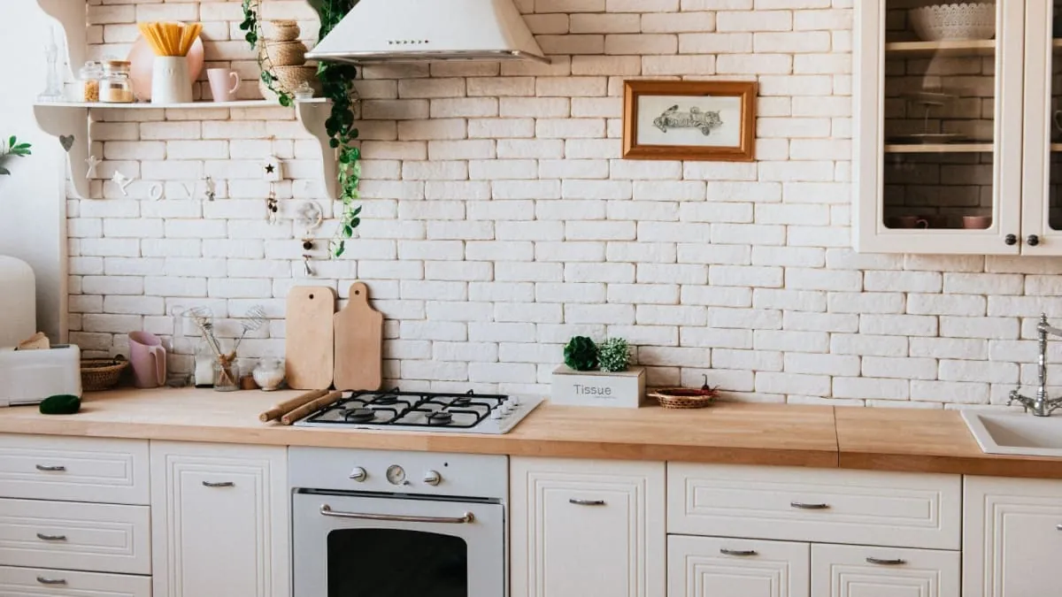 White kitchen with a brick wall, light wooden countertops, and white cabinets and appliances. A framed picture and greenery decorate the space.