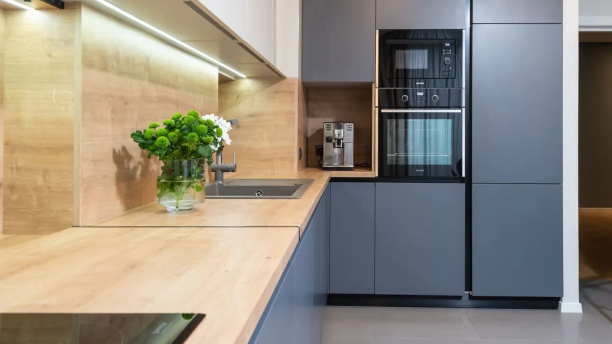 Modern kitchen with gray cabinets, light wood countertops, built-in oven and microwave, and a coffee machine.