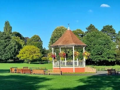 Bandstand in a sunny park, surrounded by green grass, trees, and benches.