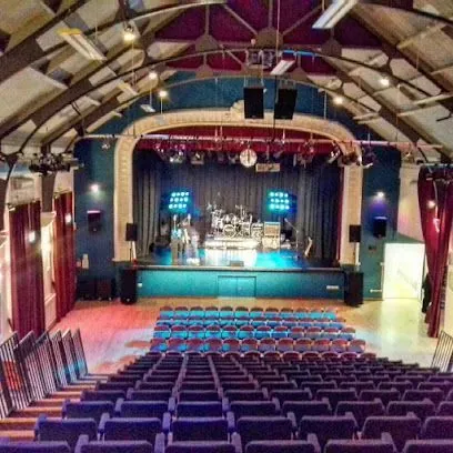 An auditorium with a stage set up for a performance, blue seating, and dark walls.