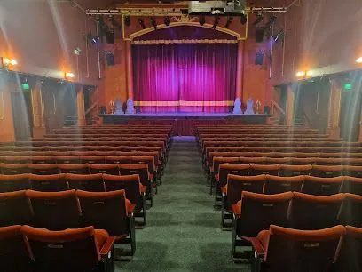 Audience view of an empty theater with red seats, stage with purple curtain, and stage lights.