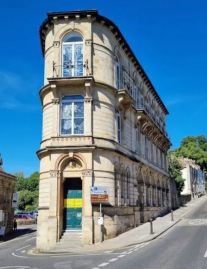 Beige building with windows, flower baskets, and a tree on a cobblestone street. A bus is visible in the background.
