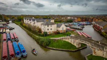 Boats moored in a canal, with houses, bridge, and cloudy sky overhead.